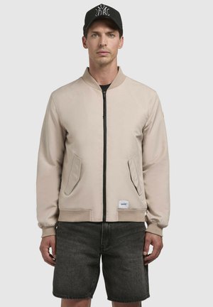 Beige Bomberjacke mit einem Frontreißverschluss, seitlichen Taschen, gerippten Bündchen und Saum. Trägt dunkle Jeansshorts und eine schwarze Kappe mit einem Logo.