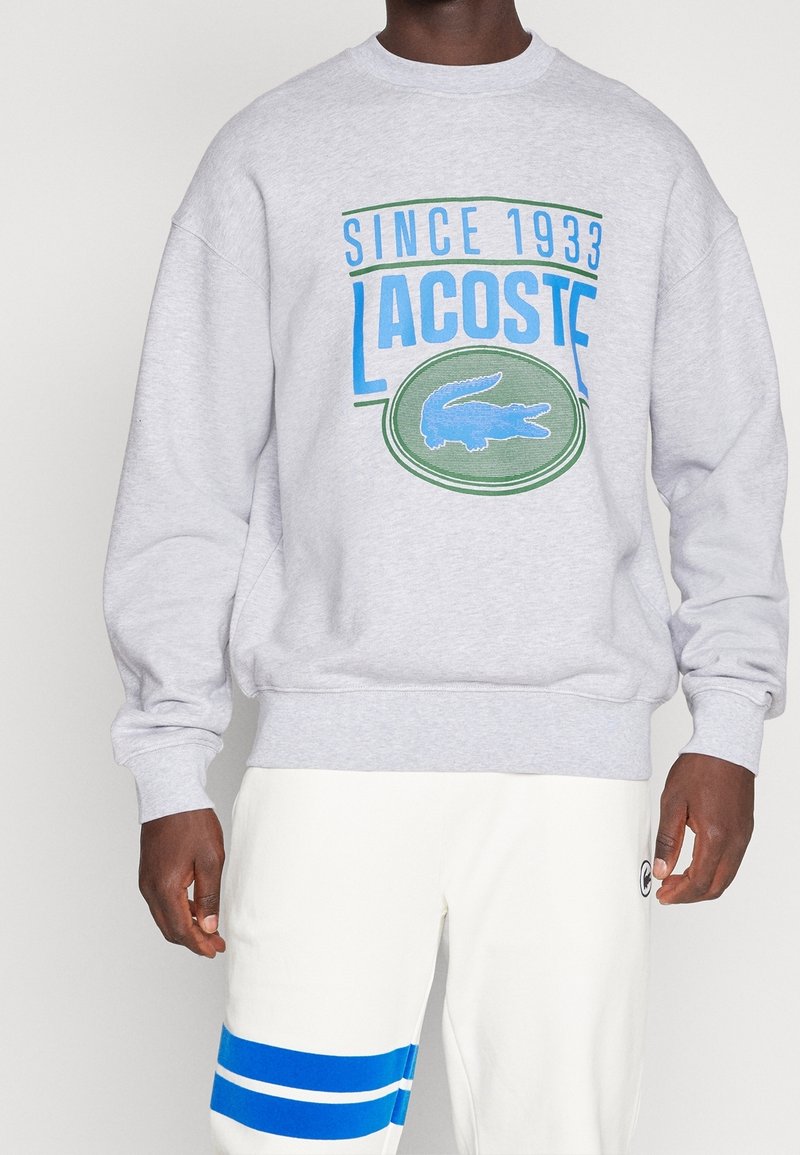 Felpa grigia con grafica blu e verde "LACOSTE" e logo di coccodrillo, abbinata a pantaloni bianchi con righe blu.