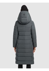 Personne portant un long manteau gris matelassé avec une capuche et un bonnet noir en tricot, vu de dos sur un fond uni.