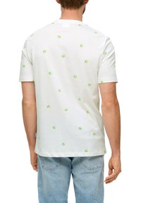 Biała bawełniana koszulka z zielonym wzorem w kształcie promieni, okrągły dekolt, krótkie rękawy i proste wykończenie. Nosi do niej jasnoniebieskie dżinsy.