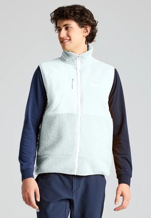 Un homme souriant porte un gilet polaire blanc zippé sur un haut bleu marine à manches longues et un pantalon, devant un fond clair uni.