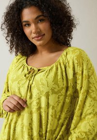 Limegrön texturerad blus med blommönster; har en rundad halsringning med en knytdetalj och långa, voluminösa ärmar.