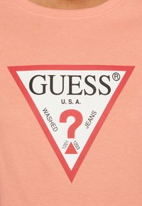 Ferskenfarvet bomulds-T-shirt med et stort trekantet logo i hvidt og rødt. Teksten inkluderer "GUESS," "WASHED," og "JEANS" med et centralt spørgsmålstegn.