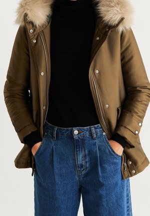Veste d’hiver marron avec capuche en fourrure portée sur un pull noir et un jean bleu taille haute, mains dans les poches.