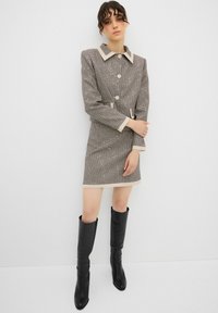 Vestido a cuadros en marrón y beige con cuello y botones, presenta un diseño ajustado y mangas largas, combinado con botas negras hasta la rodilla.