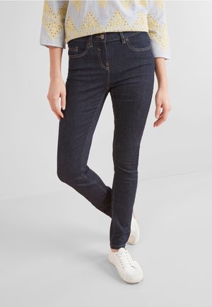 Jean slim fit bleu foncé porté par une personne avec un haut blanc et jaune à motif et des baskets blanches, debout avec une jambe croisée.