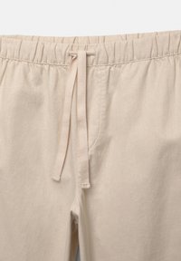 Pantalon beige avec taille élastique, cordon de serrage assorti et surpiqûres visibles sur la couture avant.
