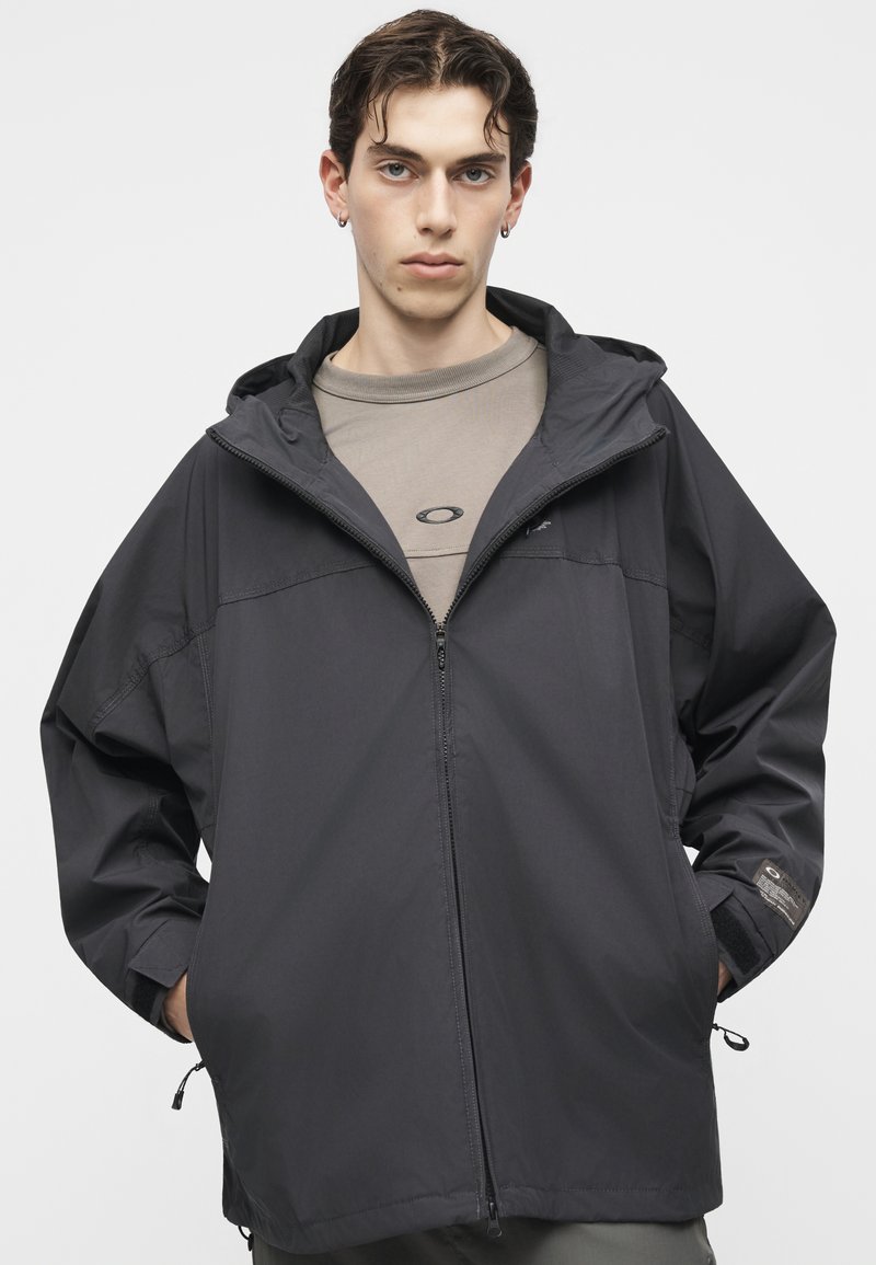 Joven con pelo corto y oscuro que lleva una chaqueta gris con capucha y cremallera sobre una camiseta topo con un pequeño logo, manos en los bolsillos, fondo neutro.