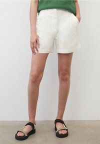 White cotton shorts with a flat front, side pockets, and a clean hem. Paired with black and olive sandals featuring a strappy design.