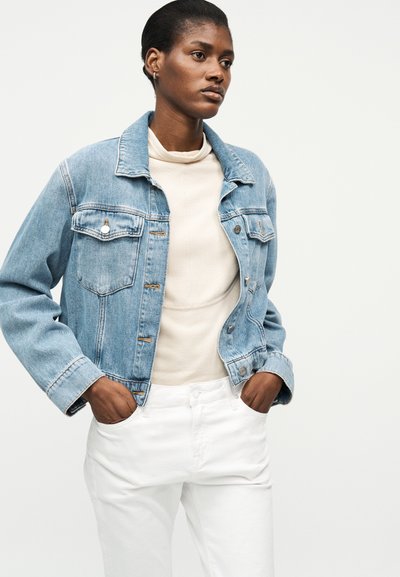 Γυναίκα με ανοιχτόχρωμο τζιν μπουφάν πάνω από μπεζ τοπ και άσπρο παντελόνι, με τα χέρια στις τσέπες, στέκεται μπροστά σε απλό ανοιχτό φόντο.