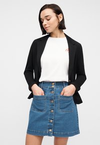 Femme portant un blazer noir, une chemise blanche avec un texte rouge, et une jupe en jean bleue à boutons sur le devant avec de grandes poches avant, les mains dans les poches, regardant vers le bas.