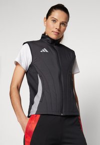 Černá bezrukávová vesta s vysokým límcem, s šedými bočními panely a předním zipem. Na horní levé straně je šedé logo Adidas.