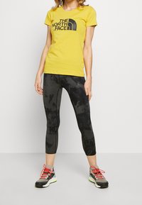 Żółta koszulka z krótkim rękawem z czarnym logo, w połączeniu z czarnymi legginsami w wzory i szarymi butami sportowymi z czerwonymi akcentami.