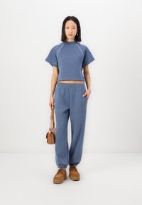 T-shirt blu corto con maniche corte, abbinato a pantaloni tuta blu coordinati. Entrambi i capi presentano un tessuto morbido e una vestibilità comoda. Borsa marrone e scarpe con zeppa.