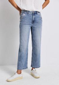 Jeans de mezclilla anchos de color azul claro con bolsillos delanteros y detalles en botón plateado. Combinados con zapatillas blancas que tienen una suela texturizada.