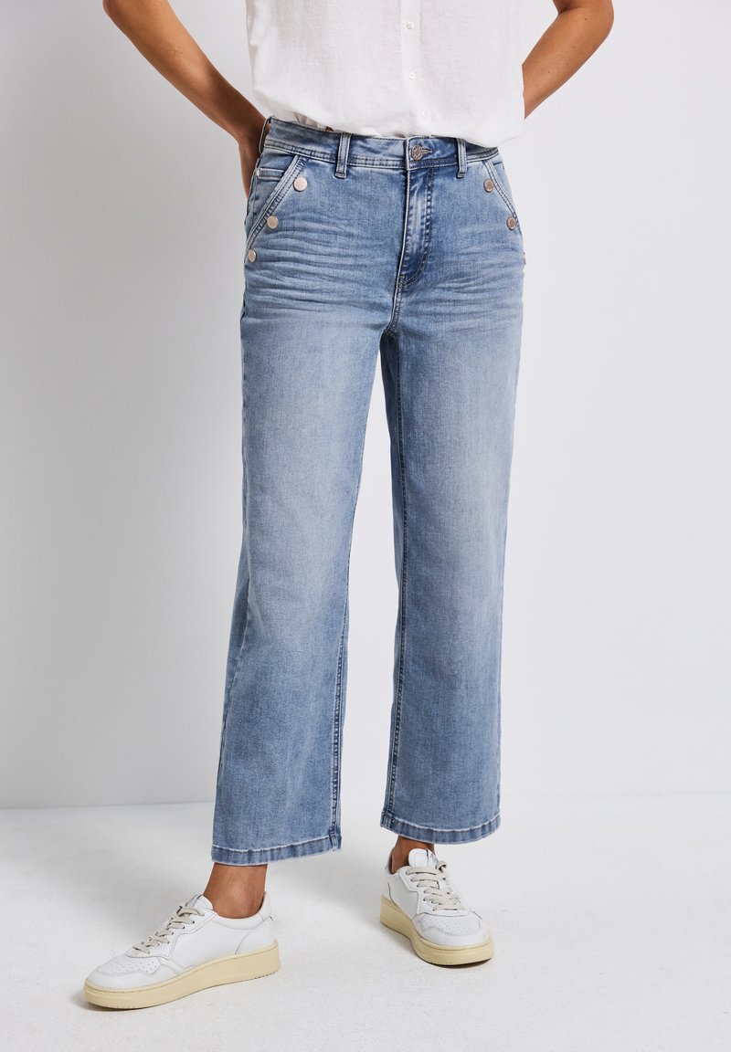 Jeans de mezclilla anchos de color azul claro con bolsillos delanteros y detalles en botón plateado. Combinados con zapatillas blancas que tienen una suela texturizada.