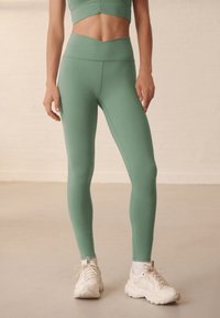 Hooggetailleerde, nauwsluitende legging in muntgroene stof met een gladde textuur. Gecombineerd met witte sportschoenen met een getextureerde zool.