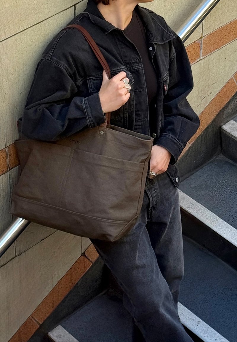 Sac fourre-tout en toile marron avec un design robuste, équipé de deux poignées supérieures et de poches avant, assorti à une tenue décontractée en denim noir.