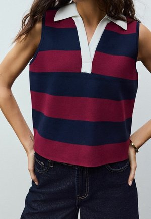 Kobieta ubrana w bezrękawnik w paski w kolorze granatowym i bordowym z białym kołnierzykiem, w połączeniu z ciemnymi jeansami, ręce spoczywające na biodrach.