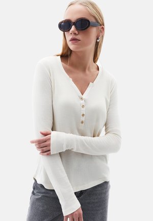 Camisa de manga larga color crema con escote en V, tapeta de cinco botones, tejido texturizado y puños elásticos. Se lleva con gafas de sol oscuras.