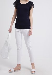 Marinblå kortärmad topp med genomskinliga axeldetaljer, parat med vita skinny jeans och vita sandaler. Modellen håller en vit tote-väska.