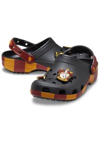 Zuecos de resina negra con acentos en franjas rojas y amarillas, que presentan un emblema de Gryffindor y agujeros de ventilación. Incluyen una correa trasera.