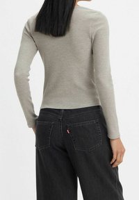 Haut à manches longues gris clair côtelé, associé à un jean noir taille haute avec poches arrière et détail emblématique de l'onglet rouge.