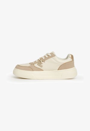 Zapatilla de perfil bajo en beige y crema con cierre de cordones, suela gruesa y charm metálico con el logo "M" en el lateral.