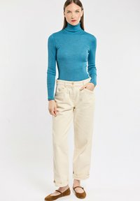 Türkiser gerippter Rollkragenpullover, crèmefarbene, locker sitzende Hose mit umgeschlagenen Säumen und braune Slippers. Einfaches, lässiges Outfit.