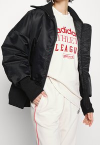 Zwarte bomberjack met een glanzende afwerking, geribbelde manchetten en een ritssluiting. Draagt het over een witte sweatshirt met rode tekst. Lichtgekleurde broek.