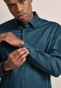 Teal Hemd mit strukturiertem Kragen und langen Ärmeln. Es hat Knopfmanschetten und eine subtile Textur, die detaillierte Nähte zeigt.