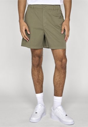 Mand iført olivengrønne shorts, hvide ankelsokker og hvide sneakers, stående mod en ensartet lys grå baggrund.