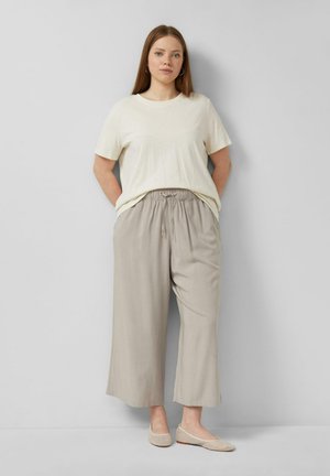 Frau steht vor einer grauen Wand, trägt ein cremefarbenes Kurzarmshirt, hellbeige, weit geschnittene Hosen und beigefarbene Ballerinas.