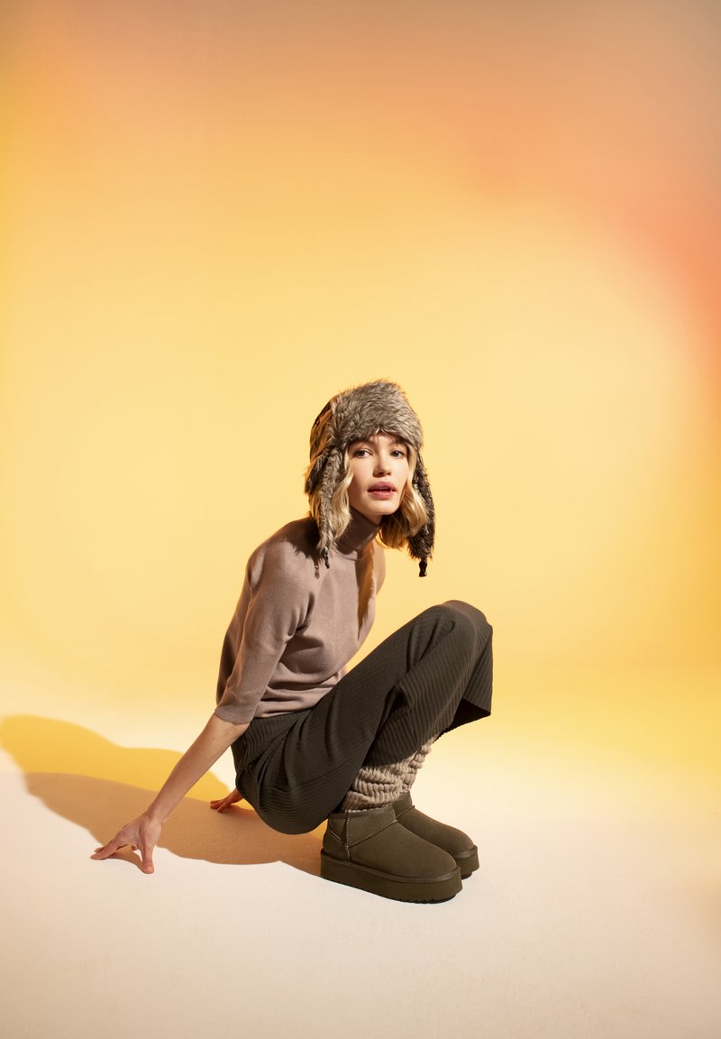 Brown fur-lined hat, light beige sweater, black ribbed trousers, green platform boots. Set against a gradient yellow to orange background.