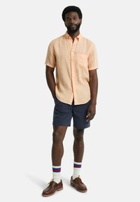 Helles orange Hemd mit kurzen Ärmeln und Brusttasche, kombiniert mit marineblauen Shorts, weißen Socken mit roten und blauen Streifen sowie braunen Schuhen.