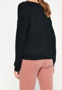 Svart ribbad stickad tröja med långa ärmar, rundad halsringning och avslappnad passform, matchad med ljusrosa kordflorsbyxor.