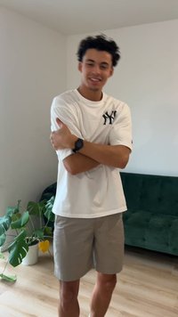 Balti marškinėliai su juodu logotipu, pilkos šortai, stovintis sukryžiuotomis rankomis. Žalios augalas ir tamsiai žalia sofa fone. Neutralios sienos.