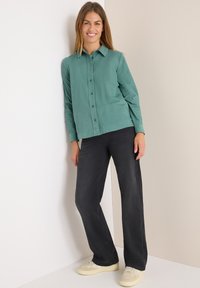 Chemise boutonnière à manches longues turquoise avec col, associée à un pantalon noir à jambe droite et des baskets claires sur un fond neutre.