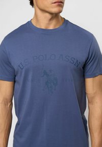 Tamsiai mėlyna medvilninė marškinėliai su apvaliu iškirpimu, dekoruoti subtiliu ekrano spaudu su polo žaidėju ir tekstu "U.S. POLO ASSN." nuo 1890 metų.