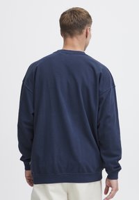 Navy plavi dukserica s dugim rukavima, okruglim izrezom i rebrastim manžetama. Mekana pamučna tkanina sa opuštenim krojem i minimalnim detaljima šivanja.