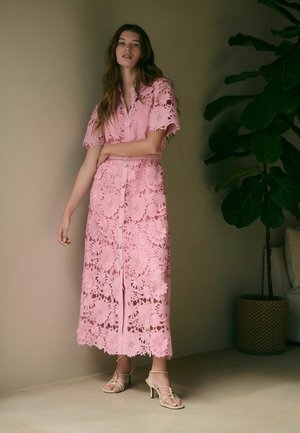 Femme portant une robe midi en dentelle florale rose avec des boutons, debout près d'un mur et d'une grande plante en pot, portant des talons beiges à brides.