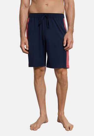 Mann trägt marineblaue Shorts mit roten Seitenstreifen, steht barfuß vor einem schlichten hellen Hintergrund.