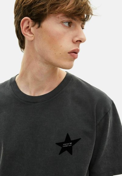 Joven con cabello corto castaño que lleva una camiseta gris oscuro con una estrella negra y un pequeño texto blanco en el pecho.