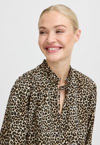 Blusa con stampa leopardata realizzata in tessuto leggero; presenta un fiocco al collo e spalle arricciate. La combinazione di colori include beige e nero.