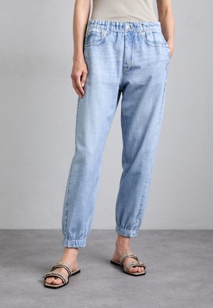 Person trägt hellblaue Jeans-Jogginghosen mit elastischem Bund und beige Schlupfsandalen, steht auf einem grauen Boden vor neutralem Wandhintergrund.