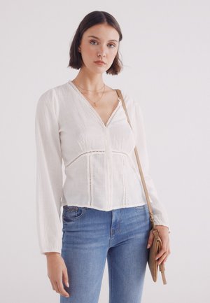 Witte, geweven blouse met lange mouwen, V-hals en knoopsluiting aan de voorkant, verfraaid met een kantafwerking. G gedragen met blauwe jeans en een kleine tas.