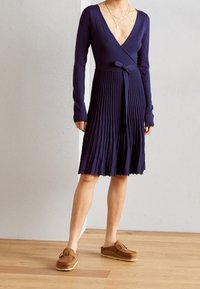 Marinblå veckad omslagsklänning med lång ärm, insnörd midja med knyt, djup V-ringning, kombinerad med bruna slip-on skor. Slät stickad textur.