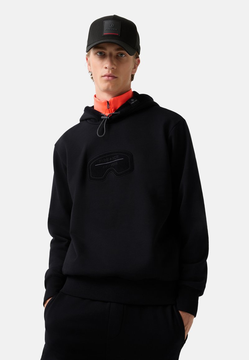 Czarna bluza z kapturem z fakturowanym, wypukłym wzorem gogli oraz sznurkiem w kapturze; w połączeniu z pomarańczowym golfem na zamek pod spodem. Czarna czapka z logo.