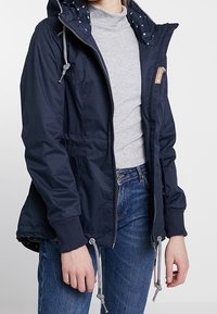Navy blue, waterproof jacket with drawstring hood, zippered front, elastic cuffs, and polka dot lining. Features a logo patch on the chest.
