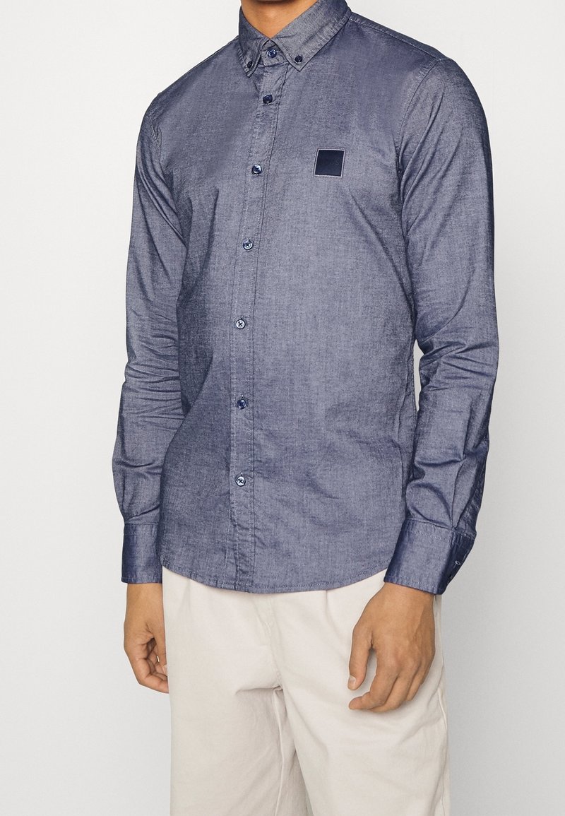Homme portant une chemise bleue à manches longues avec un petit écusson noir sur la poitrine et un pantalon beige clair sur un fond uni.